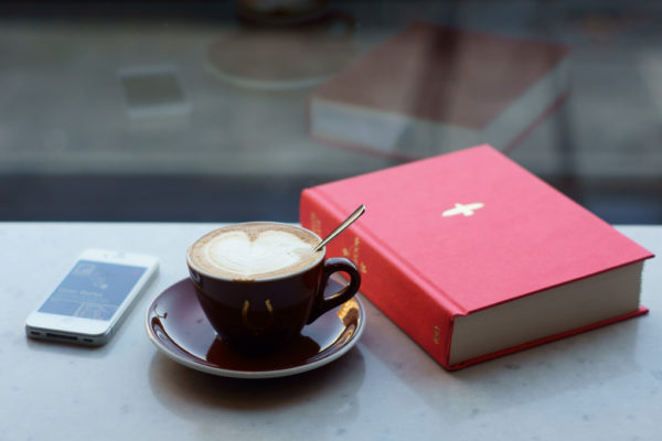 A cup of coffee with a foam heart sits in between a red hardcover book and a white smartphone.