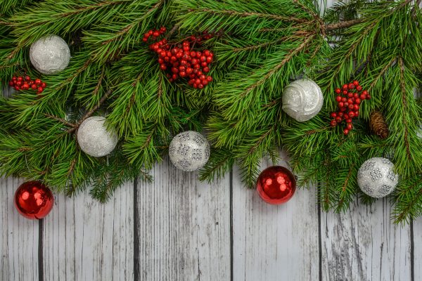 Bright red and glittery silver ornaments are nestled within evergreen garlands alongside springs of holly. The background is made up of vertical slats of weathered gray wood. Image via Pixabay.
