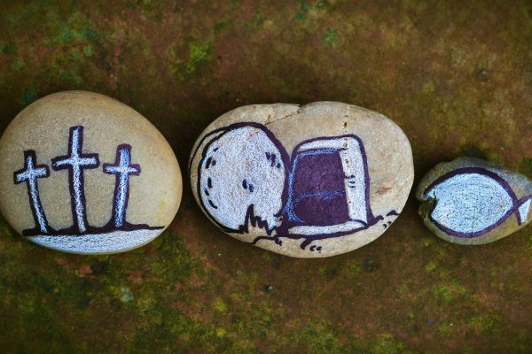 Three round tan stones sit side by side against a brown background. The first has three crosses on it, painted in white. The second depicts a white stone rolled away from a purple tomb. The third stone is much smaller than the first two and depicts a white fish.