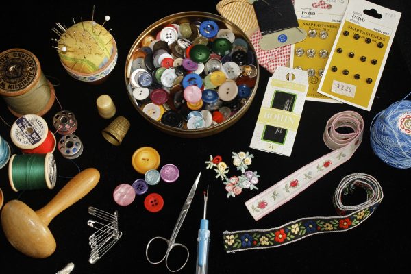 As assortment of sewing supplies is laid out against a black background. A large bowl of multicolored buttons is surrounded by thread, scissors, ribbon, a seam ripper, pins, and other bits and bobs. Image via Pixabay.