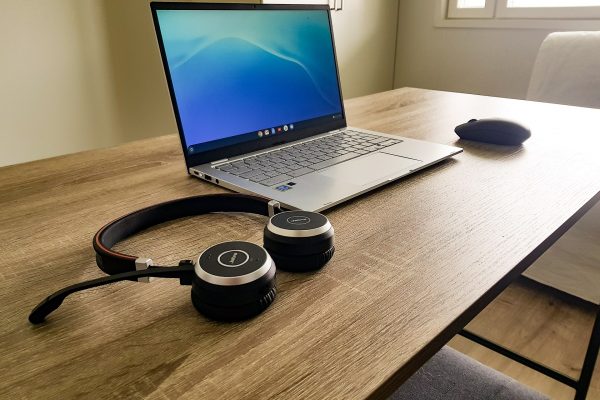 A wireless headset and mouse sit on either side of a laptop on a long wooden table.