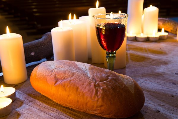 A loaf of bread sits beside a glass of wine on a wooden table, with several lit white candles in the background.