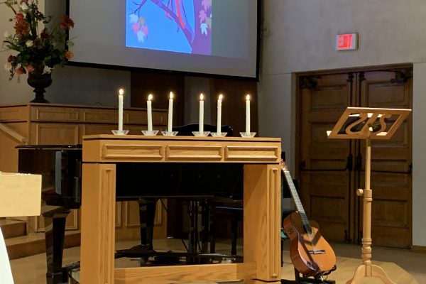 An image of the FCC Pomona Sanctuary from the 2019 All Saints' Service. 6 lit taper candles sit on a table in front of the piano.