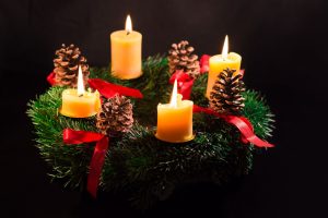 A green evergreen Advent wreath, adorned with red ribbons, four pinecones, and four lit pillar candles.