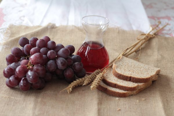 From left to right, a bunch of purple grapes, a chalice of wine, several stalks of grain, and 3 slices of bread sit on a burlap tablecloth.