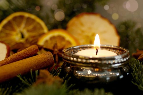 A single lit tealight sits among evergreen branches, cinnamon sticks, slices of orange, and star anise.