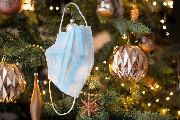 A blue disposable face mask hangs by an earloop on a Christmas tree, alongside golden ornaments and garlands.