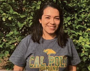 A smiling Liliana, with shoulder-length dark hair and a short-sleeve, gray, green, and yellow Cal Poly Pomona t-shirt, stands in front on a leafy green shrub.