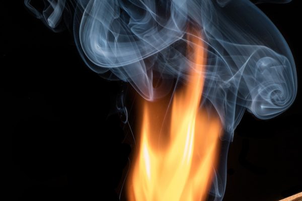 A single lit match burns brightly against a black background while gray smoke rises upward. Image via Pixabay.