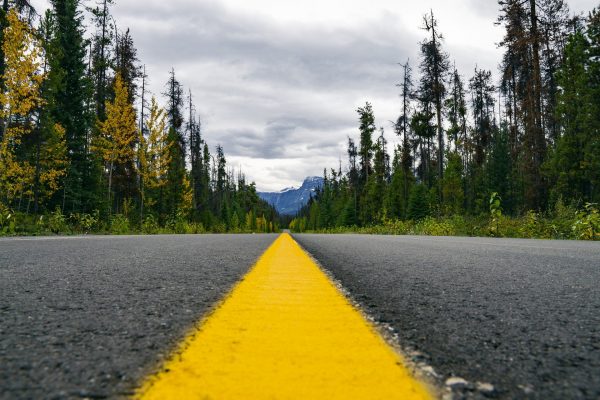 A close-up of a bright yellow line running down the center of an asphalt road, lined by trees on either side. A mountain peak is visible in the distance. Image by Joshua Woroniecki from Pixabay.