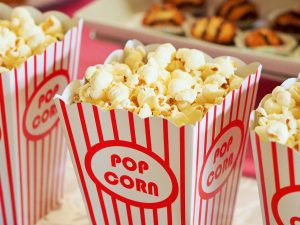 3 red and white striped containers of popcorn sit alongside each other in a row amongst other snacks. Image by Devon Breen from Pixabay.