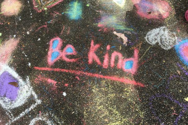 The words, "Be Kind" are written in pink chalk on black asphalt. Other chalk drawings surround the words as beams of sunlight stream in from outside the frame. The gaps in the b, e, and d of "Be kind" are filled in with bright blue chalk. Image via Pixabay.
