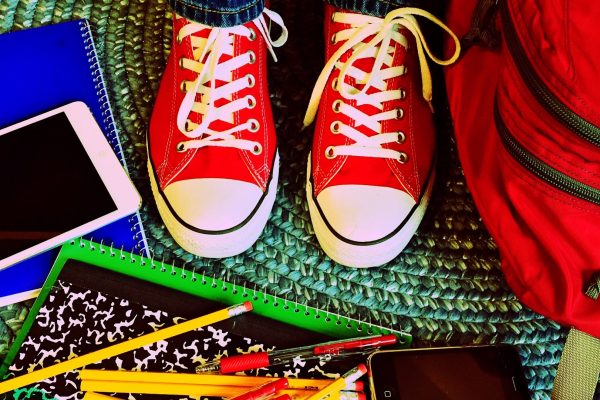 A pair of red high top shoes sit amidst scattered school supplies--including a red backpack, numerous pens and pencils, notebooks, a phone, and a tablet--on a green-blue braided rug. Image via Pixabay.
