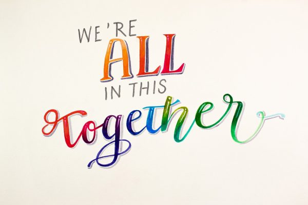 The words, "We're ALL in this together," are written across a cream-white background. The words "ALL" and "together" are written in rainbow colors; "together" is also written in cursive. Image via Pixabay.