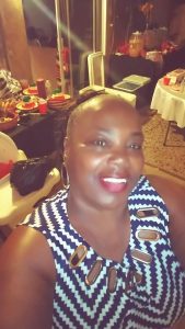 Natalie Ford-Brown, wearing a blue and white chevron-striped tank top with cut out detailing, smiles into the camera. She is also wearing red lipstick. Tables of food and drinks are set up in the background.