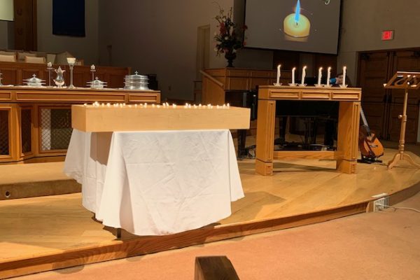 A wooden box of lit candles sits atop a table covered in a white cloth in the center of the FCC Pomona sanctuary stage. Six tall taper candles are lit on an uncovered wooden table to the right of the box of candles. The screen behind the piano shows a single lit candle under the words, "All Saints' Sunday".