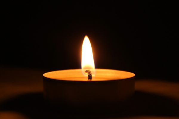 A single lit tea light. The candle sits in a circular metal tin; the wick burns brightly against the dark background. The tin's shadow is visible around its periphery. Image via Pixabay.