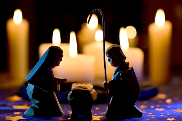 Dark carved wooden figures of Mary and Joseph sit on either side of a manger. Joseph holds a tall shepherd's crook. The figures are situated atop a blue tablecloth with gold stars, and numerous lit candles of various shapes and heights are visible in the background.