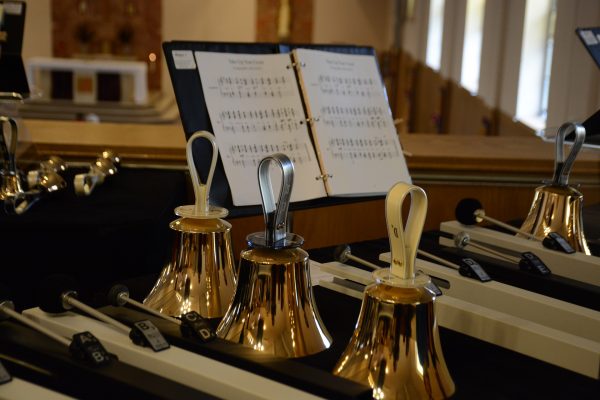 A vertical row of three shiny handbells sits in between vertical rows of mallets of various sizes. A black binder filled with sheet music is propped up on a stand behind the bells. The table is lined with black cloth. Other bells of various sizes are visible around the central row. Image by JenniferWebb from Pixabay.