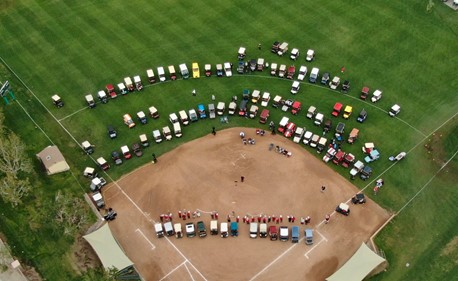 An aerial view of several rows of golf carts fan out around a softball diamond. One row of golf carts spans the diamond; two other rows face the first from across the green field. Tiny figures of people are visible on both the diamond and the field.
