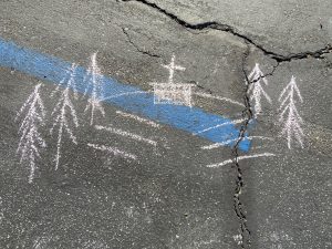 A white chalk drawing of several benches in front of a central rectangle with a cross in the center. Tall trees line the sides of the hill. The drawing is drawn on black asphalt; the blue line of an accessible parking space bisects the drawing.