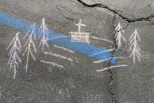 A white chalk drawing of several benches in front of a central rectangle with a cross in the center. Tall trees line the sides of the hill. The drawing is drawn on black asphalt; the blue line of an accessible parking space bisects the drawing.