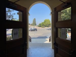 One set of the dark brown wooden doors of the church are open onto the parking lot. One of the archways of the church's portico is framed on either side by one of the doors. It is a sunny and clear day outside; green trees are visible in the distance.