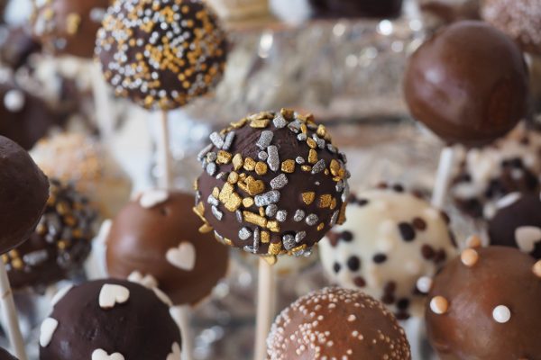 An assortment of white, milk, and dark chocolate cake pops. Each is covered with a variety of sprinkles, such as white hearts, gold and silver flecks, and black, brown, and white balls. Image via Pixabay.