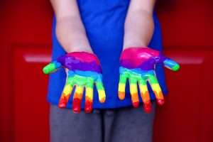 A child with white skin stands with their hands outstretched. Only a portion of their hands, arms, and lower torso are visible. Their hands are covered with bright rainbow stripes; they are wearing a blue shirt and gray pants. They appear to be standing against a red door. Image via Pixabay.