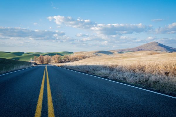 A single lane asphalt road stretches towards the horizon. The road is shaded, but the grassy and mountainous landscapes to the left and right of the road are largely illuminated by sunlight. The sky is blue and scattered with white-gray clouds. Image via Pixabay.