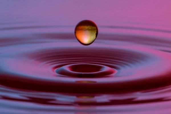 A orange-yellow drop of water creates ripple effects in a pool of purple-pink water. Image via Pixabay.