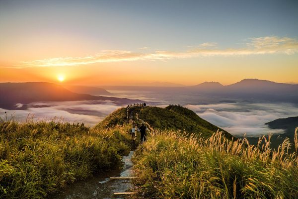 A grassy green mountain peak overlooks a sweeping vista of other distant peaks. The sun rises on the right side of the image, and white clouds swirl around the based of the mountains. A path runs down the center of the grassy peak, framed on either side by a wooden railing. Image via Pixabay.