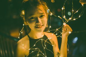 A young girl in a dark dress sits on a bench and smiles at the camera as she holds a twisty strand of fairy lights. The lights glow white-yellow in the dark.