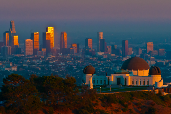 A view of the Los Angeles skyline at sunset, with Griffith Observatory in the foreground. Angled sunlight glints off of the tallest skyscrapers and the Observatory's hill. Image via Pixabay.