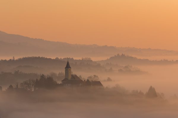 A white church steeple rises above a landscape of tree-covered hills, shrouded in early morning fog. The sky is a pale gold orange. Image via Pixabay.