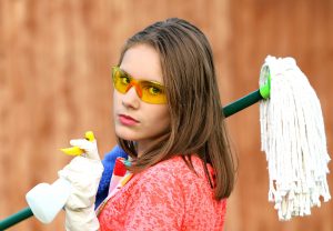 A person with shoulder-length brown hair and clad in a red-pink shirt looks over their shoulder with a serious expression. They are wearing white gloves and yellow-orange safety goggles. A mop is slung over one of their shoulders and they are holding a spray bottle. Image via Pixabay.