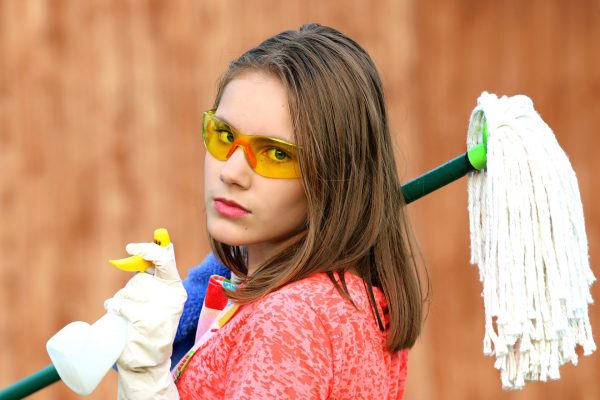 A person with shoulder-length brown hair and clad in a red-pink shirt looks over their shoulder with a serious expression. They are wearing white gloves and yellow-orange safety goggles. A mop is slung over one of their shoulders and they are holding a spray bottle. Image via Pixabay.