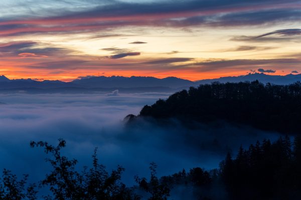 Tree-covered hills rise above the morning fog. The sky is streaked with red, blue, and white clouds. Image via Pixabay.