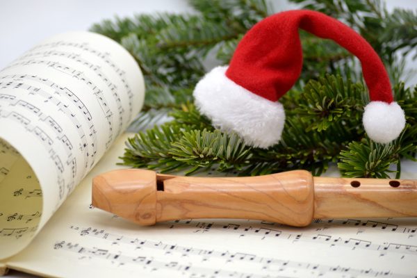 A light wooden recorder sits atop a book of music. A small Santa hat sits atop an evergreen sprig. Image via Pixabay.
