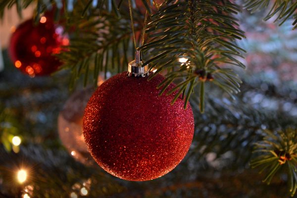 A sparkly red spherical Christmas ornament sits in the center of the image. It is hanging amidst the evergreen branches and twinkling lights of a Christmas tree. Other red ornaments are visible in the background.
