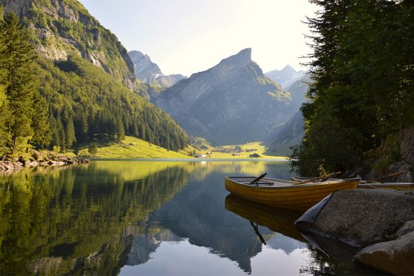 Several canoes rest against the rocky shore of a still body of water. The green mountains and trees that line the banks are reflected in the water's surface. It is a sunny and clear day. Image via Pixabay.