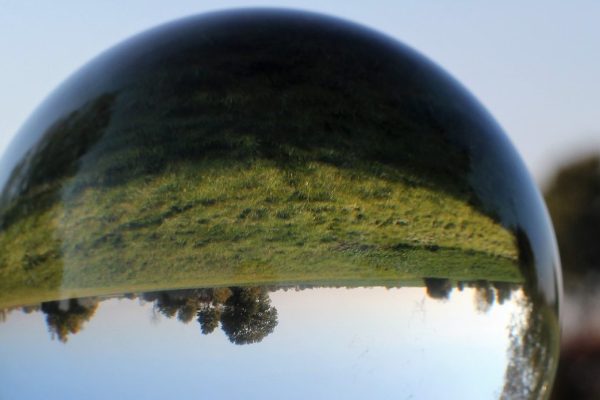 A clear crystal sphere sits on a stand, showing the inverse of the landscape in the background: A grassy green field with distant trees. Image via Pixabay.