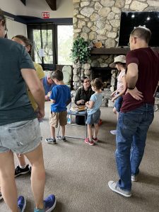 Church members gather around a mini Communion table inside a building at the Loch Leven Camp and Retreat Center. A woman with pale skin, dark glasses, a black jacket, and with her brown hair tied back, kneels beside a blue plastic chair atop which the Communion elements sit: King's Hawaiian rolls and a paper cup of juice. Several children, seen from the side, process past this station to receive their Communion elements; other adults are seen from behind.