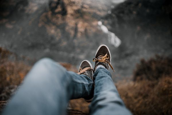 Two legs, clad in light-wash denim pants, hang over a rocky ledge. This lower half of a person also wears dark brown sneakers with light brown laces. The vista below is blurry, but appears to be some sort of forested mountain area, colored in shades of gray, brown, and white. Image via Pixabay.