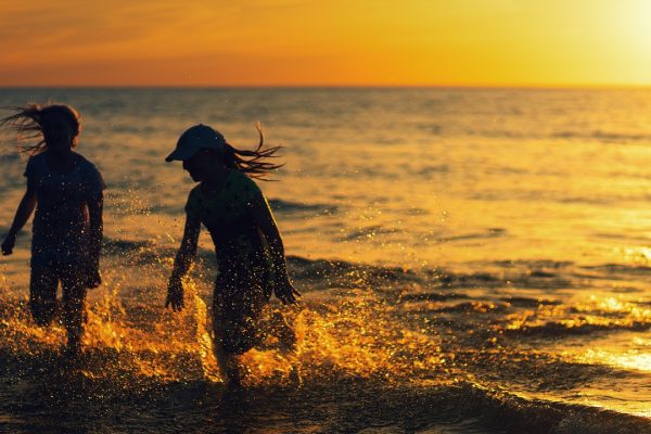 Two kids, both with long hair flying over their shoulders, splash in the ocean. Spray flies up around them. The setting sun glints off of the water to the right of the image. Both of the kids are shadowy, obscuring their features; one is wearing a baseball cap. Image via Pixabay.