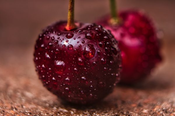 A close-up image of two deep red cherries. They have been recently washed and are covered with water droplets. The stems are a green-gold tone. It is difficult to ascertain the texture of the surface on which the cherries rest, but it is a speckled brown color. Image via Pixabay.