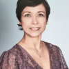 A professional headshot of Hilda Koorn, director of the Child Development Center. She is a Latina woman with short dark hair. She’s wearing a lacy short-sleeved purple blouse and pendant necklace. The background is a light gray-blue.