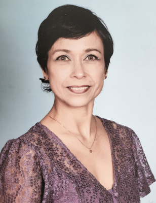 A professional headshot of Hilda Koorn, director of the Child Development Center. She is a Latina woman with short dark hair. She’s wearing a lacy short-sleeved purple blouse and pendant necklace. The background is a light gray-blue.