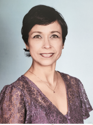 A professional headshot of Hilda Koorn, director of the Child Development Center. She is a Latina woman with short dark hair. She’s wearing a lacy short-sleeved purple blouse and pendant necklace. The background is a light gray-blue.
