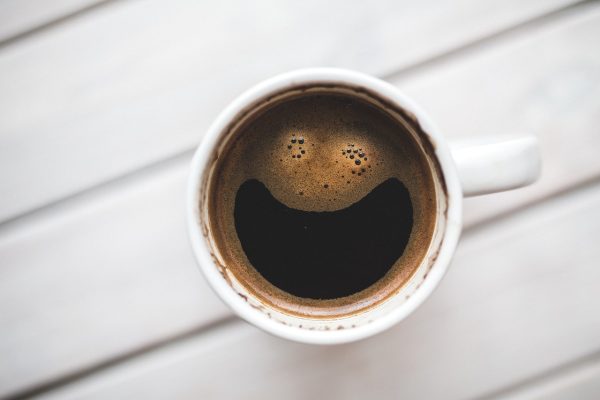 A cup of coffee in a white mug as seen from above. The bubbles on the surface of the coffee appear to be forming eyes, with a large bubble-free portion below that looks like a smiling mouth. The mug sits on a surface covered with diagonal white boards. Image via Pixabay.